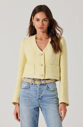 A woman is wearing a light yellow, textured, cropped blazer with long sleeves and a rounded neckline. It features three buttons down the front and two large patch pockets on the front. She pairs it with light blue high-waisted jeans and a beige belt with a gold buckle. She completes the look with gold earrings and a gold necklace.